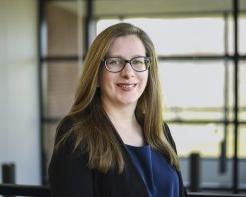 Alyssa Schleicher in a blue shirt and glasses, stands in front of windows on the Utica campus, 微笑.